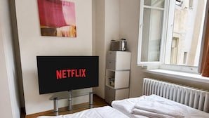 A hotel room with a bed, a TV displaying 'NETFLIX,' a radiator, and a window. - Cozy Studio in the Heart of Zurich - Stork 25 (Zürich)