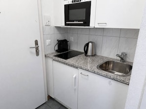Eine kompakte Küche mit weißen Schränken, einer eingebauten Mikrowelle, einem Wasserkocher und einer Spüle aus Edelstahl.