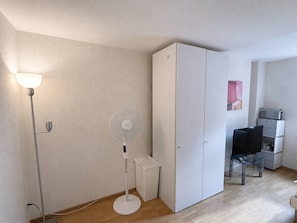 Ein Zimmer mit Standventilator, Schreibtisch, Stuhl und Kleiderschrank.