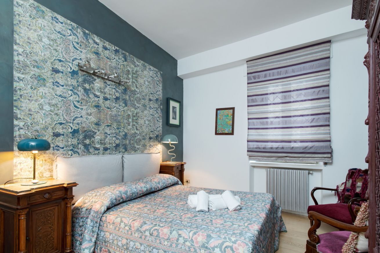 Un dormitorio con una colcha estampada, una mesita de noche con lámpara, un radiador y una ventana con persiana a rayas.