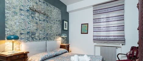 A bedroom with a patterned bedspread, a nightstand with a lamp, a radiator, and a window with a striped blind.