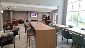 A modern dining area with wooden tables and chairs, a flat-screen TV mounted on the wall, and a view of the outdoors through large windows.