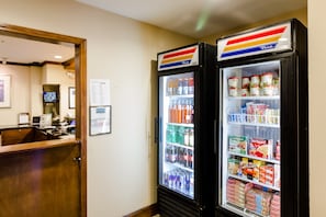 A hotel lobby with a reception desk, two True Value vending machines stocked with beverages and snacks, and a framed menu on the wall.