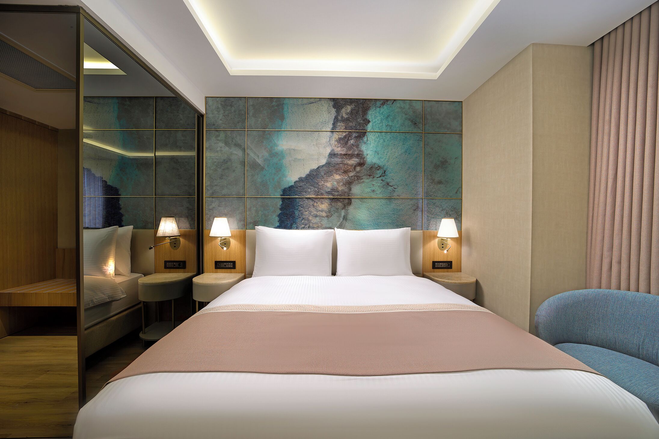Ein Hotelzimmer mit einem großen Bett, zwei Nachttischen inklusive Lampen, einer Sitzecke mit einem Sessel und einem großen, abstrakten Wandbild.