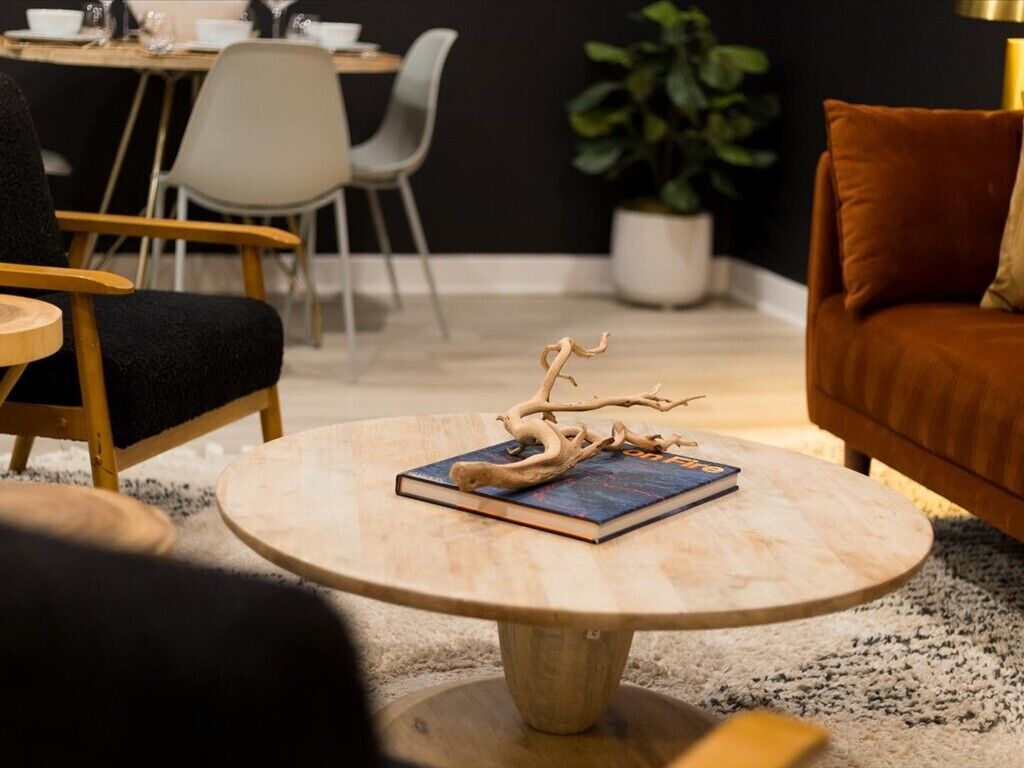 A round wooden table with a book and a branch decoration, a sofa, and a dining area in the background.