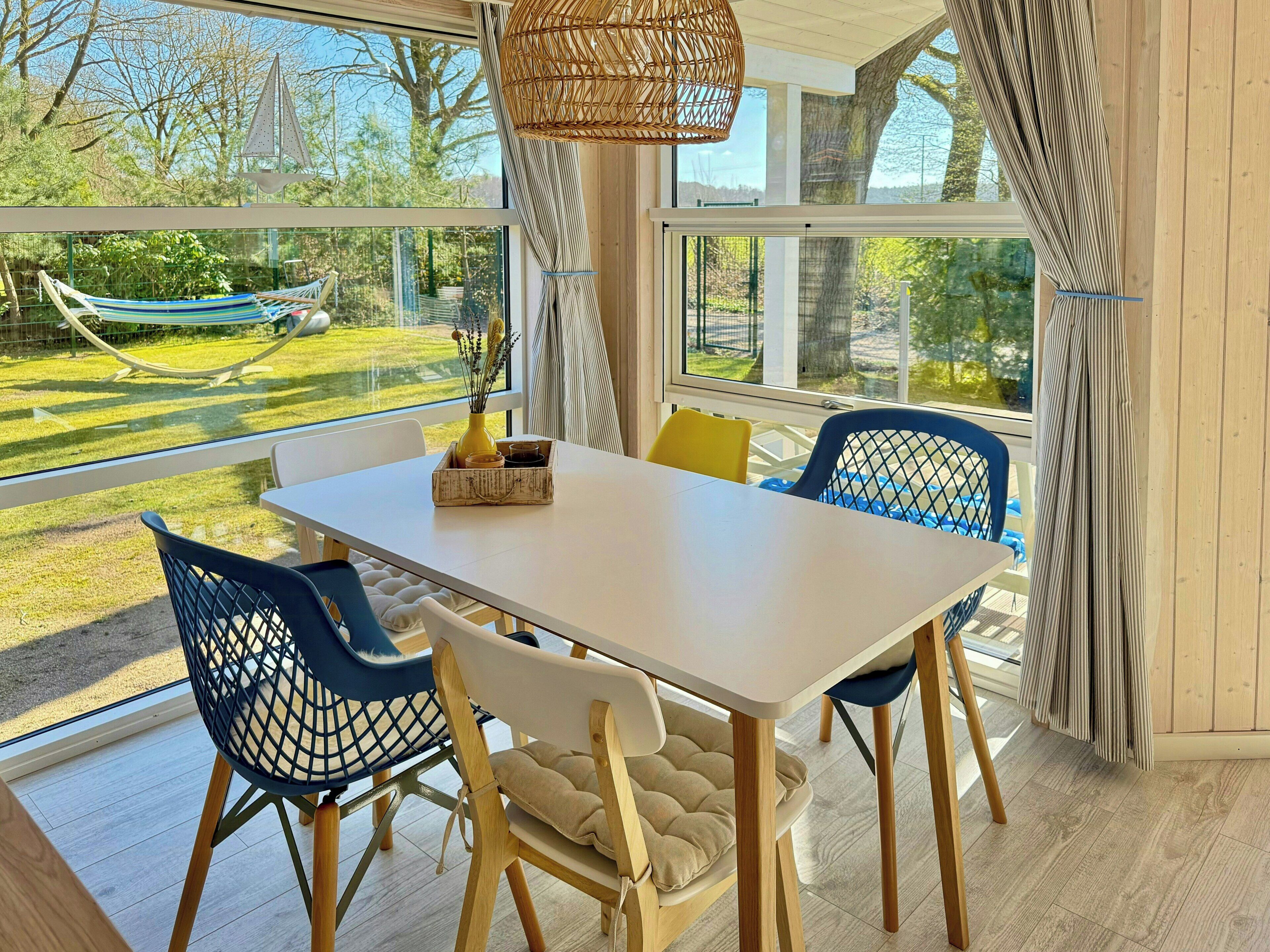 A dining area with a white table, blue and white chairs, and a view of a garden with a hammock.