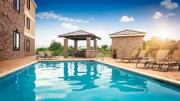 A swimming pool with lounge chairs and a gazebo, surrounded by stone walls and a building with large windows.