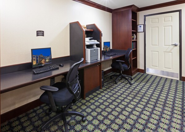 A hotel room with two desks, each with a computer, keyboard, and mouse. There is a bookshelf and a door with a sign on it.