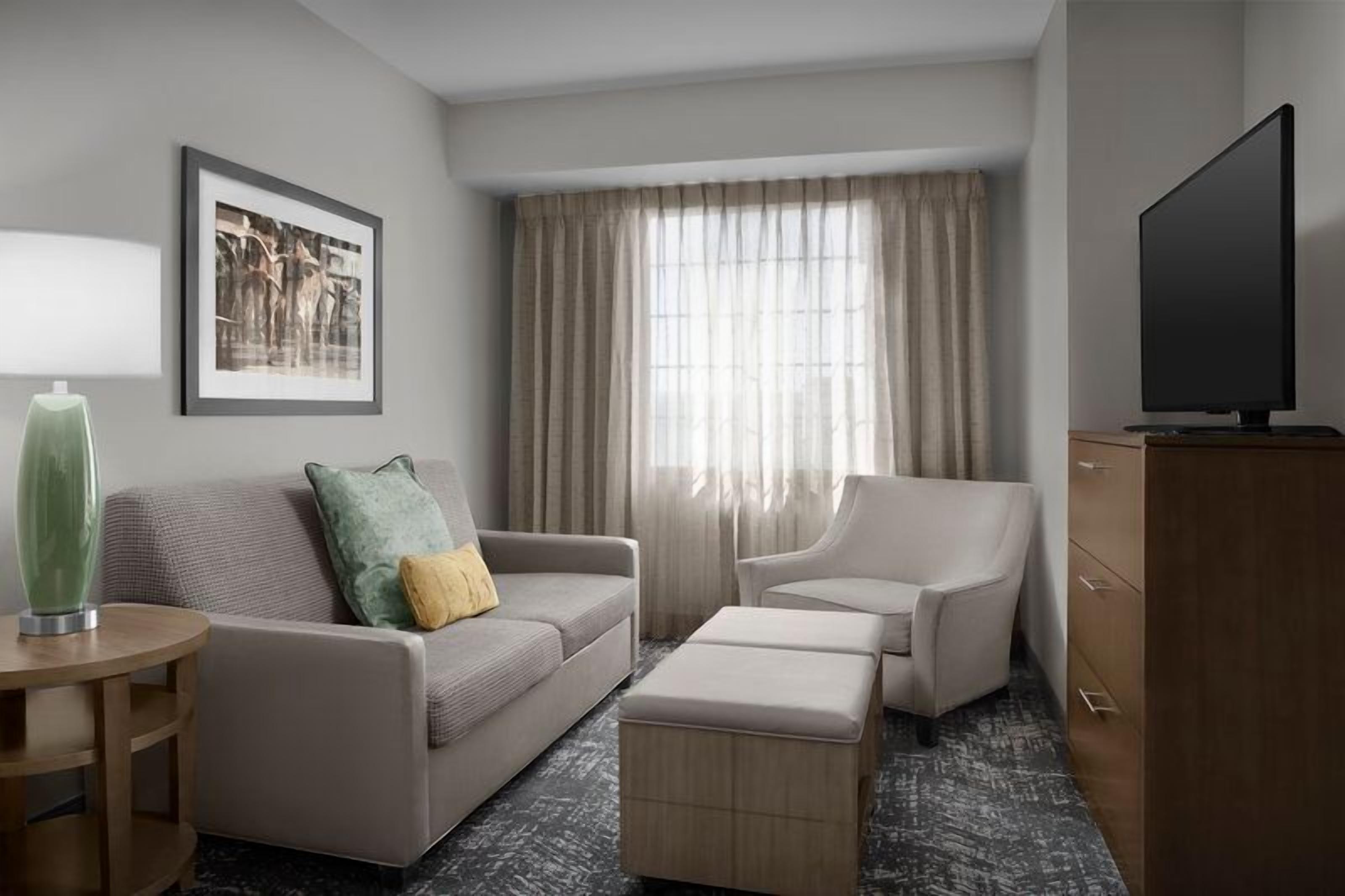 Ein Hotelzimmer mit Sofa, Ohrensessel, Hocker, Flachbildfernseher, Beistelltisch und Lampe.