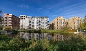 Eine Reihe moderner Apartmentgebäude mit Balkonen, ein ruhiges Gewässer im Vordergrund und ein klarer, blauer Himmel.