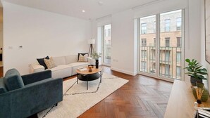Uma sala de estar moderna com um sofá, uma mesa de centro e uma poltrona azul. Há uma janela grande com vista para edifícios.