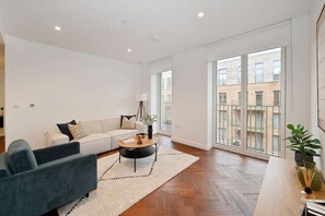 Ein modernes Wohnzimmer mit einer Couch, einem Couchtisch und einem blauen Sessel. Ein großes Fenster bietet Blick auf Gebäude.