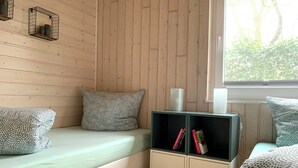 Una habitación con una cama, un pequeño aparador con libros y un ventanal con vistas a árboles.