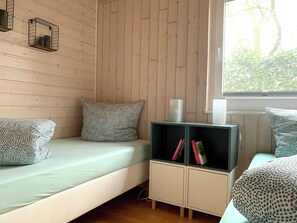 A room with a bed, a small cabinet with books, and a window with a view of trees. - Charming wooden house by the lake with sauna and boat (Dümmer)