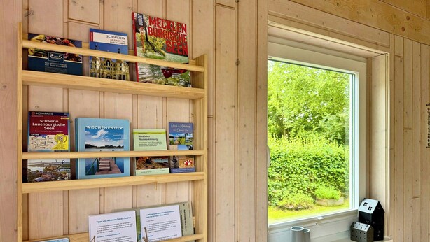 Una estantería de madera con libros de viajes y mapas, una ventana con vistas a zonas verdes y una pequeña mesa con un tapete.