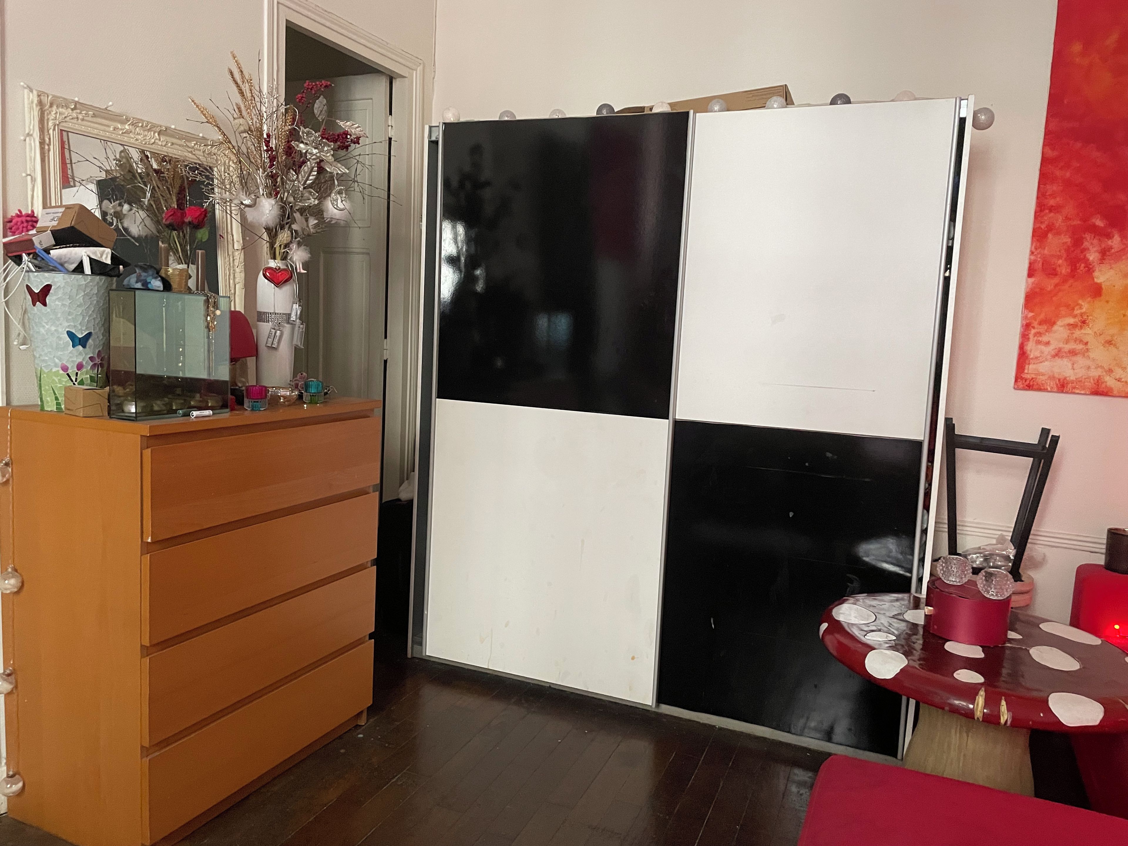 A room with a wooden dresser, a black and white checkered cabinet, a small table with a red chair, and a mirror with decorative items.