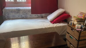 Une petite pièce avec un lit simple, une fenêtre avec des barreaux, une table de chevet et un panier en osier contenant divers objets.
