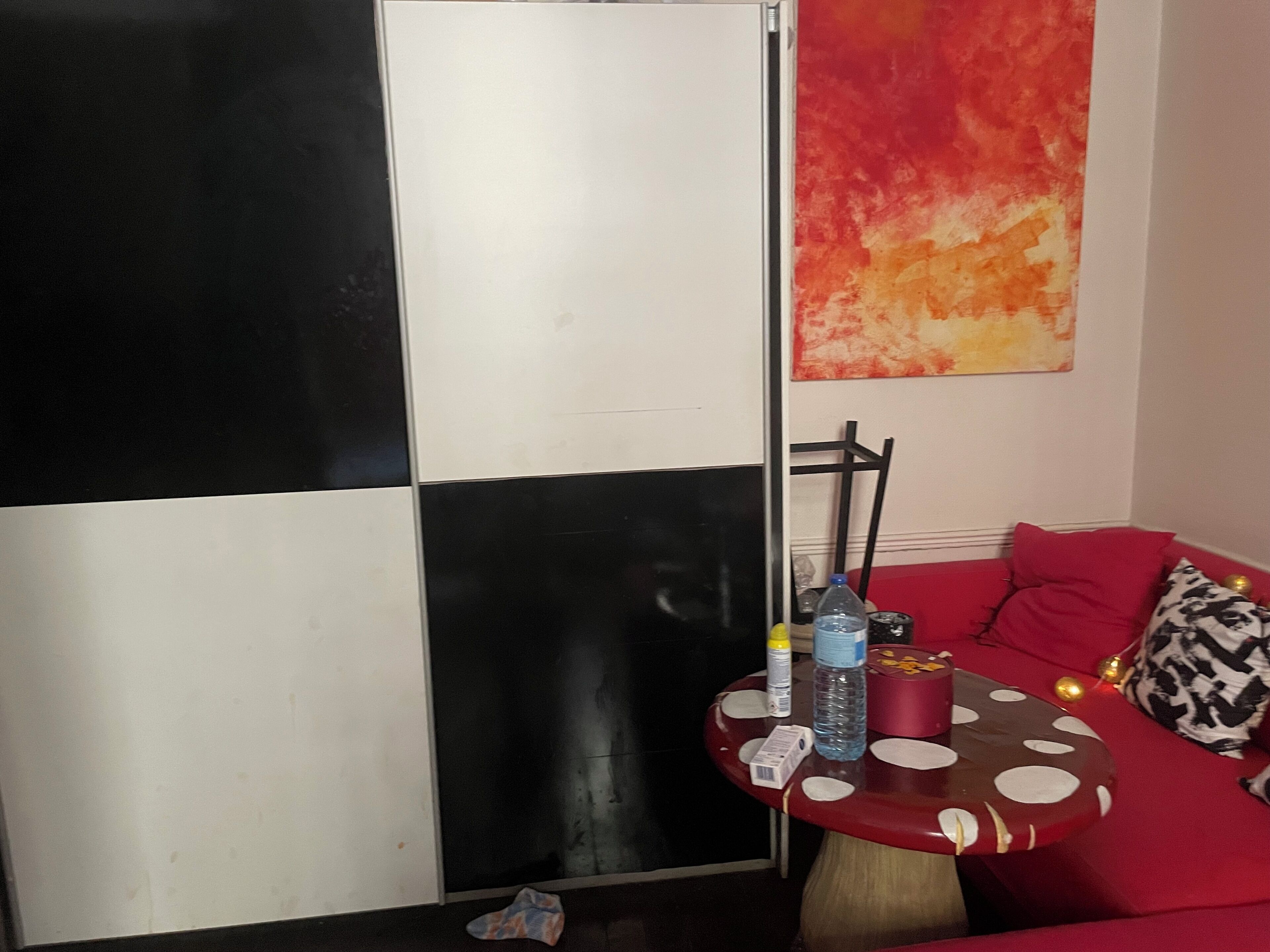 Une pièce avec un sol à carreaux, un canapé rouge, une petite table ronde recouverte d’une nappe à pois et un mur orné d’un tableau abstrait rouge.
