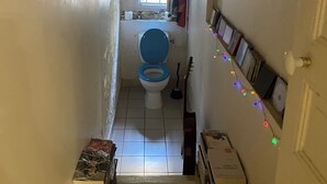 Un couloir étroit mène à une salle de bain équipée de toilettes, d’une fenêtre et d’une étagère où sont rangés des livres et un balai.