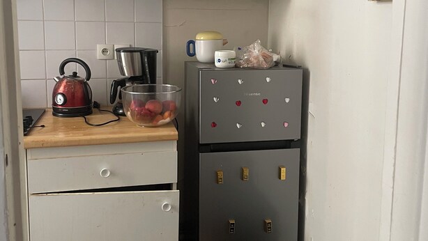 Une cuisine compacte avec des meubles blancs, une bouilloire en acier inoxydable, une machine à café et un réfrigérateur décoré d’aimants.