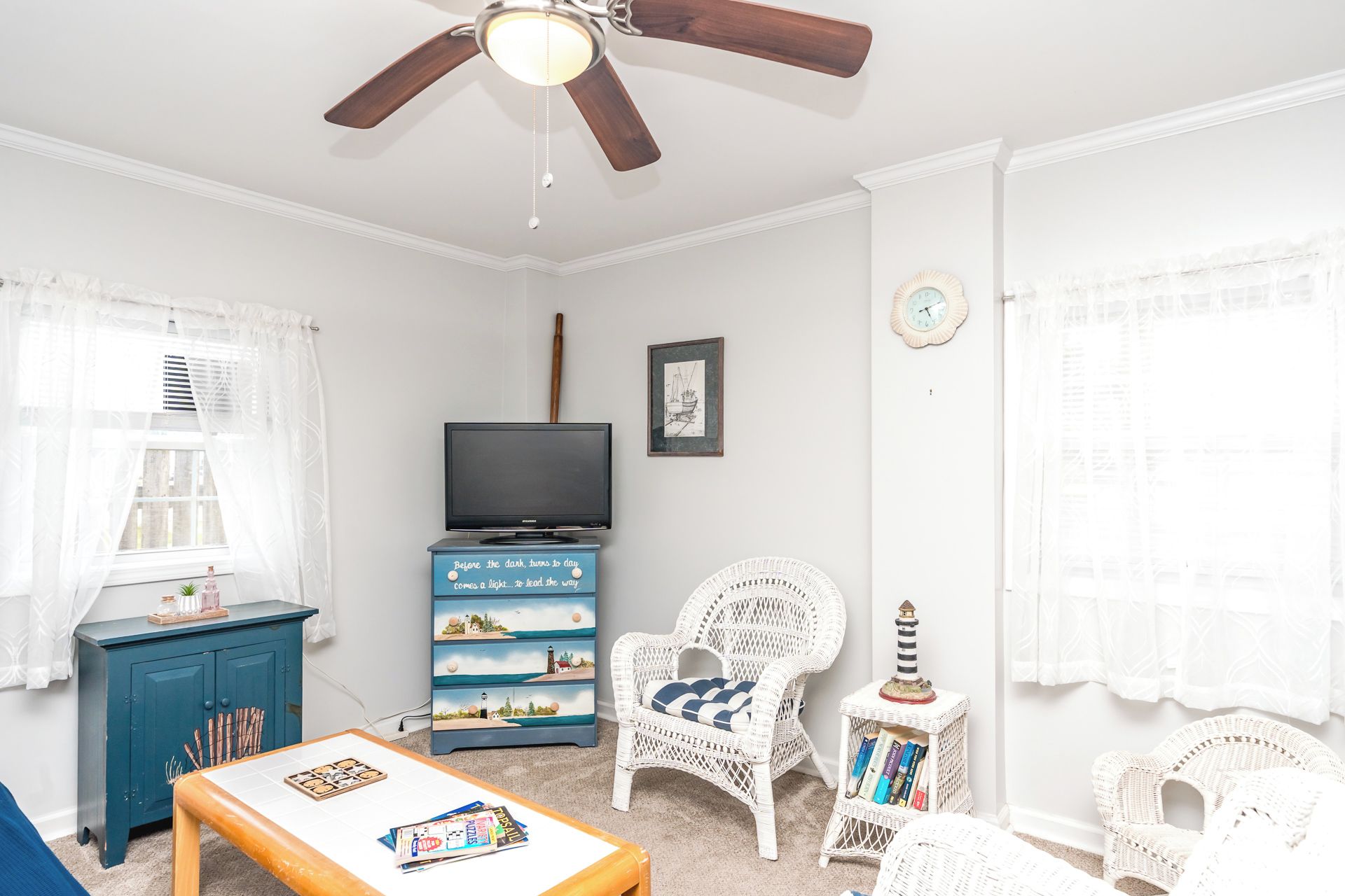 A room with a ceiling fan, a television on a blue cabinet, a wicker chair, and a small table with books.