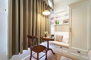 Una habitación con una silla de madera, una mesita redonda pequeña con un jarrón y un reloj, un sofá con un cojín de cuadros, y un armario.