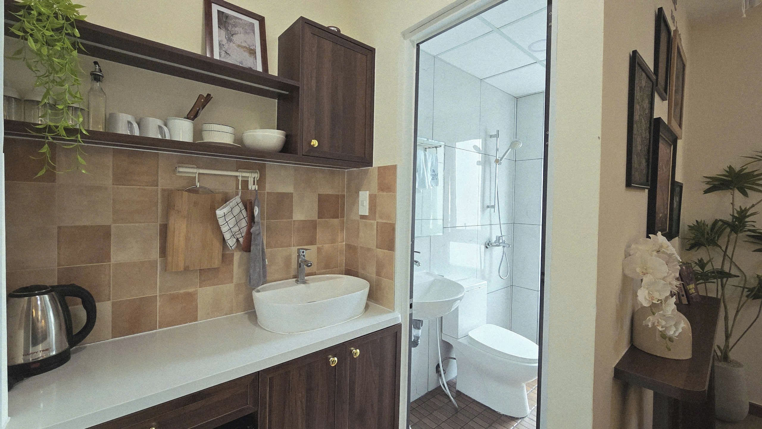 A bathroom with a large sink, a shower, and a toilet. There is a shelf with kitchen items and a plant.
