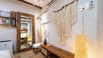 Hotelkamer met houten vloer, een bed, een bankje, een spiegel, een wandairconditioner, een macramé wanddecoratie, een vaas met takken, en een lamp.