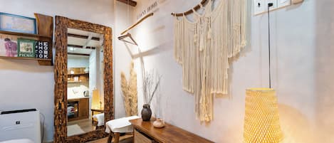 Hotelkamer met houten vloer, een bed, een bankje, een spiegel, een wandairconditioner, een macramé wanddecoratie, een vaas met takken, en een lamp.