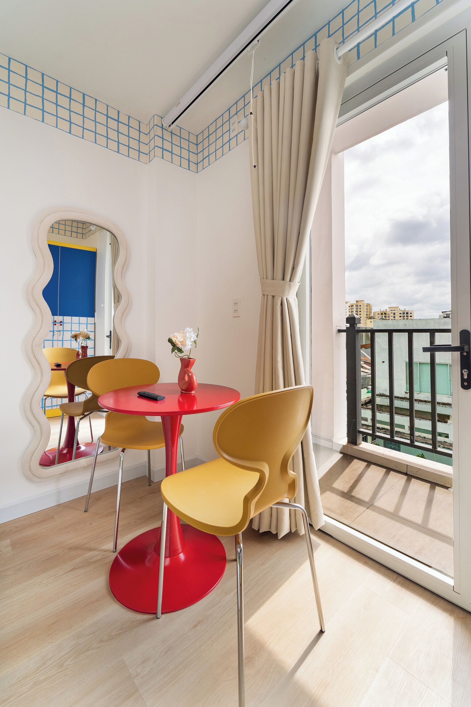 Ein modernes Zimmer mit einem roten Tisch und gelben Stühlen, einem Spiegel mit wellenförmigem Rahmen sowie Blick auf Gebäude durch eine Balkontür.
