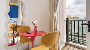 Ein modernes Zimmer mit einem roten Tisch und gelben Stühlen, einem Spiegel mit wellenförmigem Rahmen sowie Blick auf Gebäude durch eine Balkontür.
