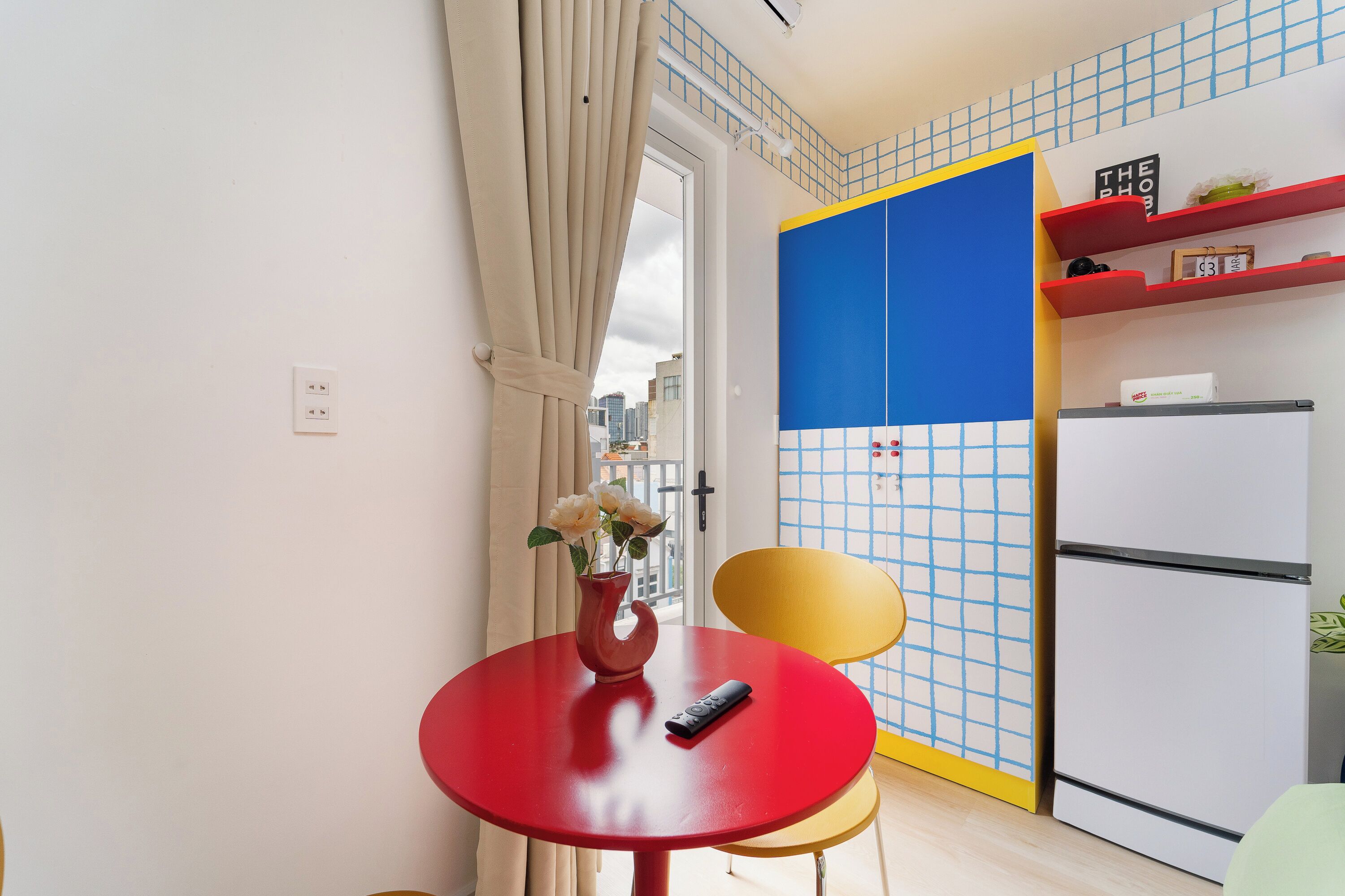 Een kleine keuken met een rode tafel, een gele stoel, een witte koelkast en een blauw-geel betegelde achterwand.