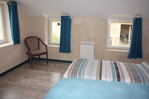 Ein Schlafzimmer mit einem Bett, einem Sessel, zwei mit Vorhängen versehenen Fenstern, einer Heizung und Blick ins Freie.