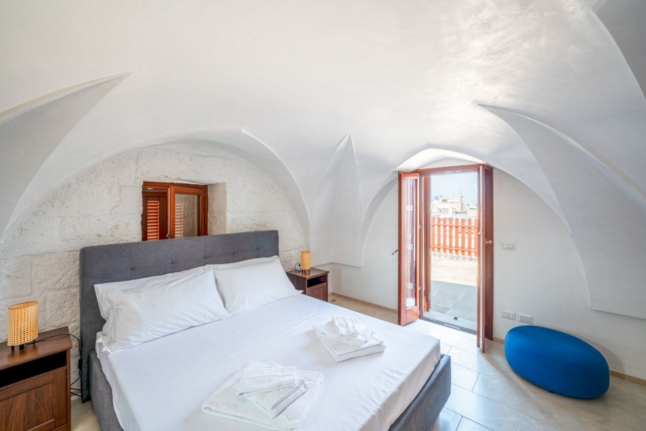 A bedroom with a bed, a nightstand, a lamp, a blue chair, and a view through an open door.