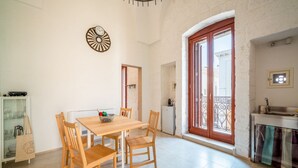 Uma sala de jantar com mesa e cadeiras de madeira, um relógio decorativo na parede e uma cozinha com janela e varanda.