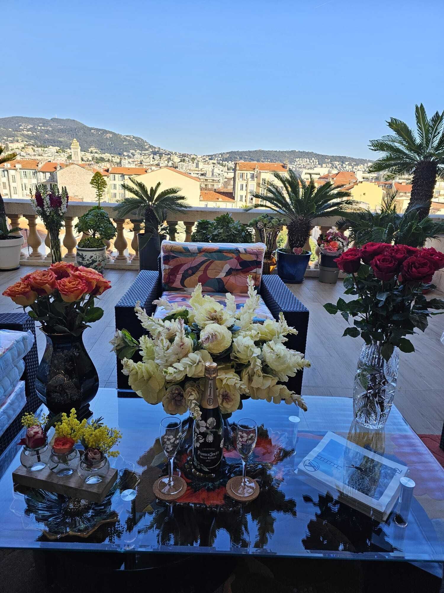 A table set with flowers, a bottle, and glasses, overlooking a cityscape with buildings and hills.
