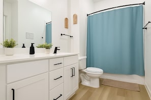 Un baño moderno con tocador blanco, pomos negros, inodoro blanco y cortina de ducha azul.