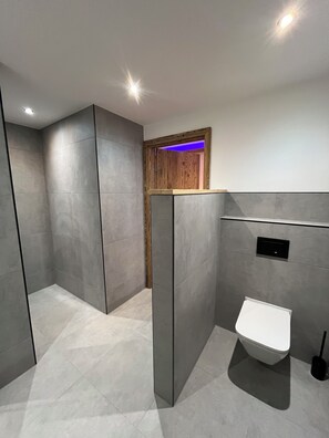 A modern bathroom with grey tiles, a wooden door, a white sink, and a black flush toilet. - Alpine luxury in the heart of the Allgäu—Alpenchic Apartments Eichten (Pfronten)