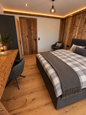 Une chambre avec un plancher en bois, un lit recouvert d’une couverture à carreaux, un bureau avec une chaise et une armoire en bois.