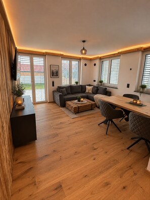 A spacious living area with a wooden floor, a dark-colored sofa, a dining table with chairs, and a ceiling light fixture. - Alpine luxury in the heart of the Allgäu—Alpenchic Apartments Eichten (Pfronten)