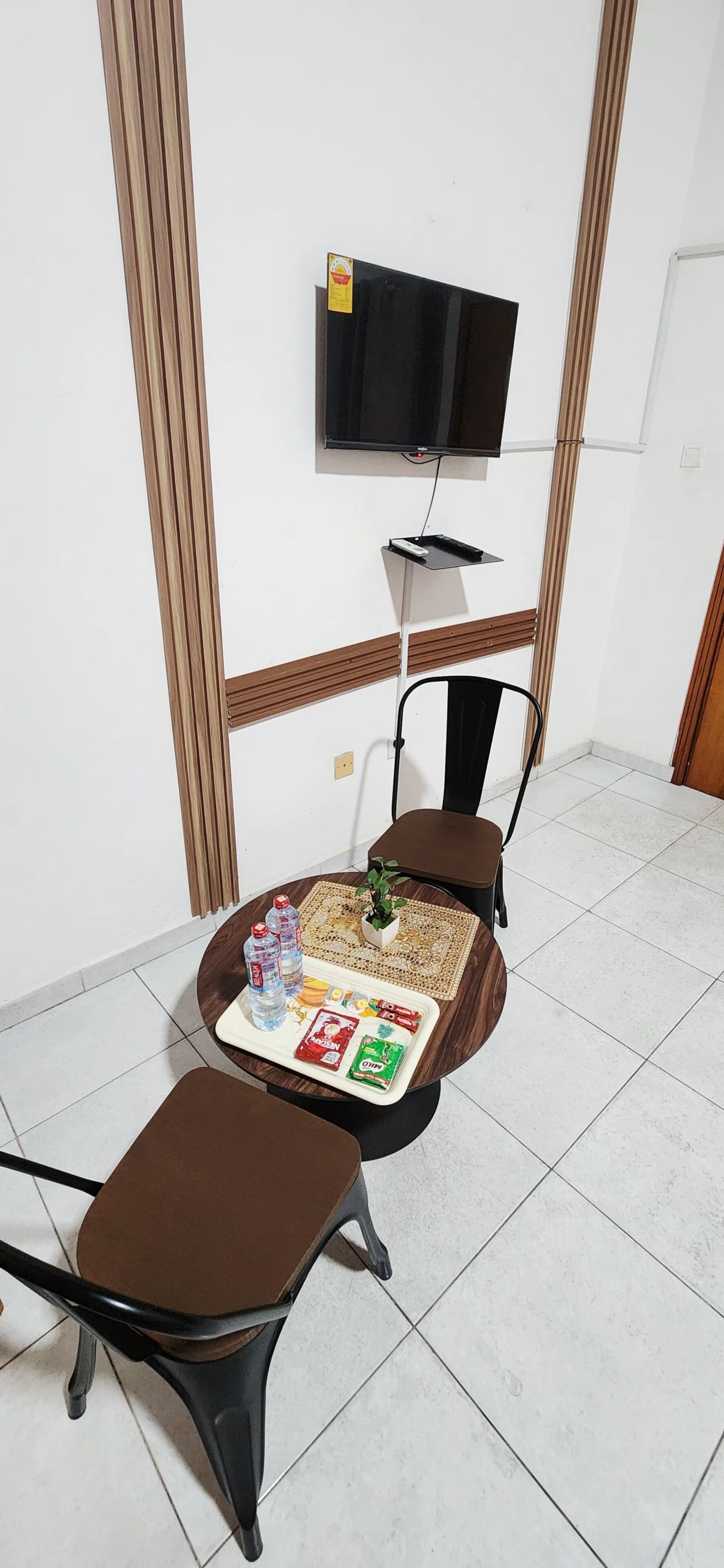 A small room with a single chair, a small table with snacks, a television mounted on the wall, and a tiled floor.