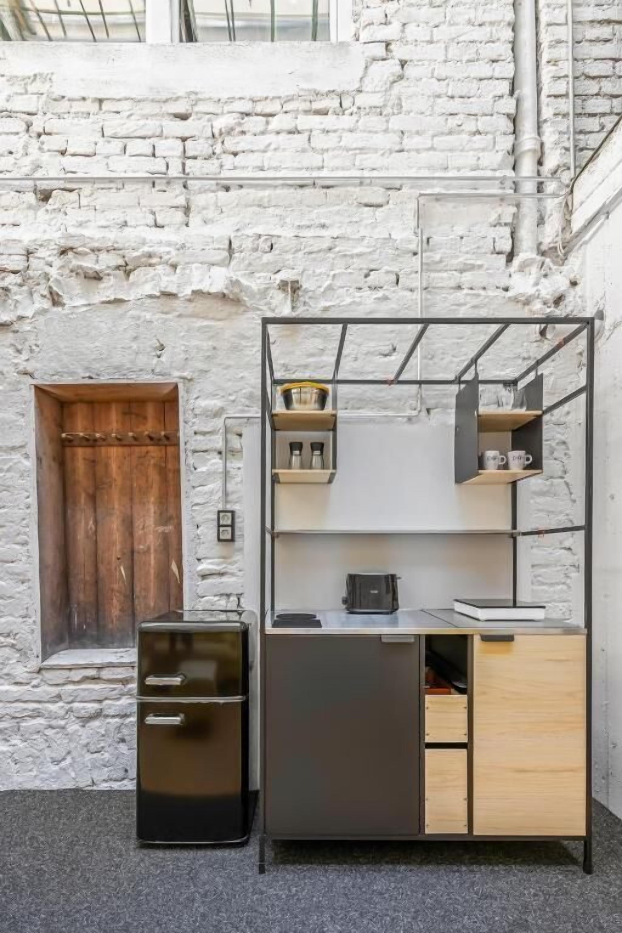 A compact kitchenette with a black refrigerator, a wooden cabinet, and a metal shelf unit against a white brick wall.