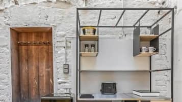 A compact kitchenette with a black refrigerator, a wooden cabinet, and a metal shelf unit against a white brick wall.