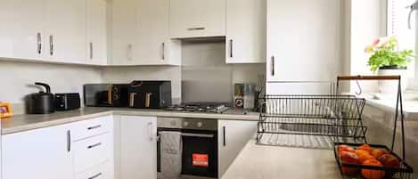 Une cuisine moderne avec des meubles blancs, une cuisinière à gaz, un four à micro-ondes et un panier de fruits.