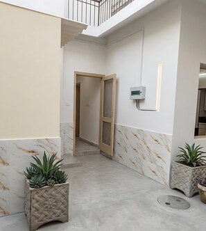 A corridor with a door leading to a room, a wall-mounted electrical box, and two potted plants. - Qaya Guest House (Islamabad)