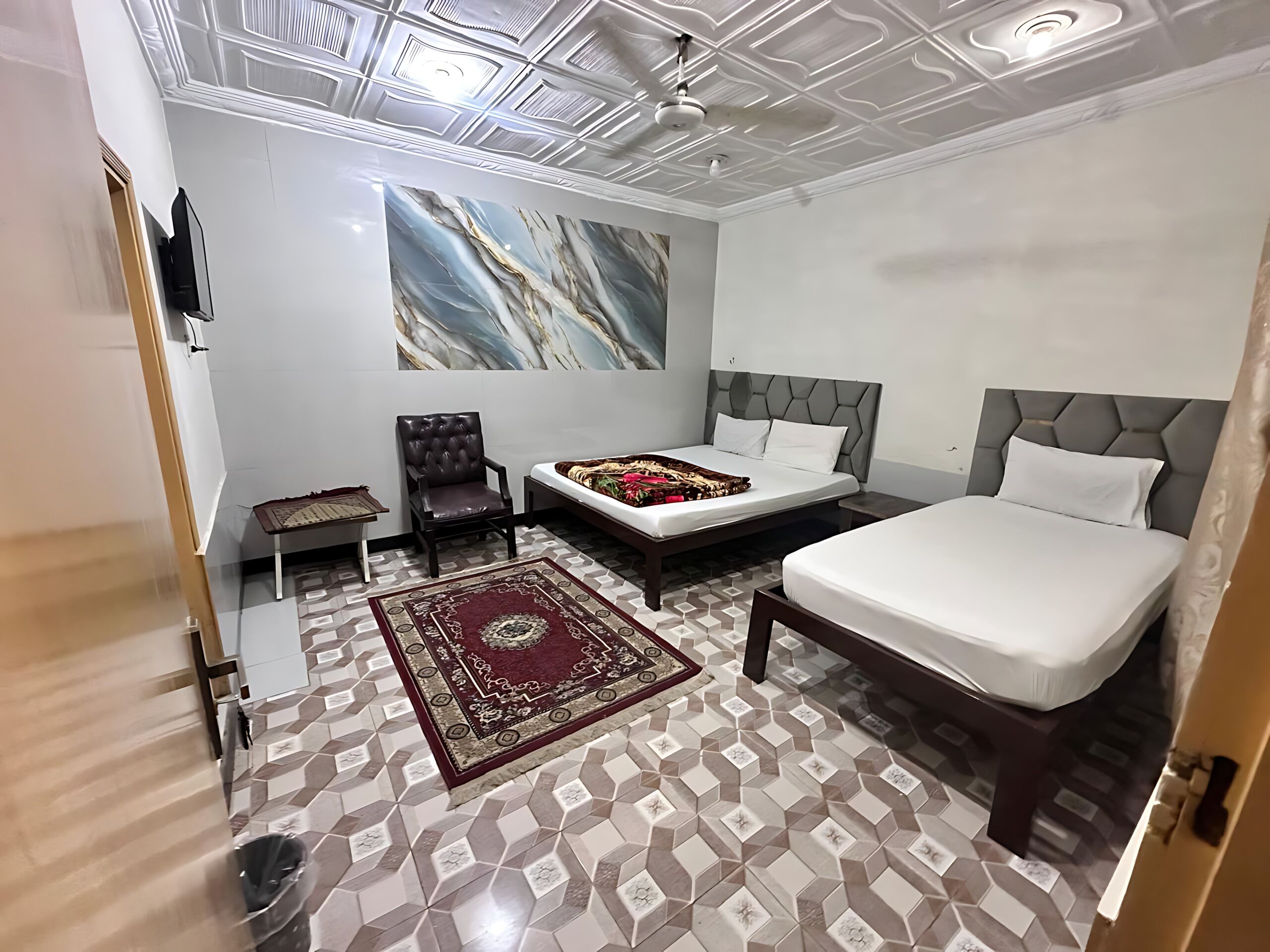 A hotel room with a bed, a chair, a small table, a decorative rug, and a ceiling with a geometric pattern.