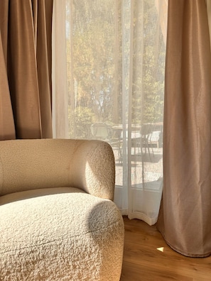 A room with a sofa, a window with sheer curtains, and a view of an outdoor area.