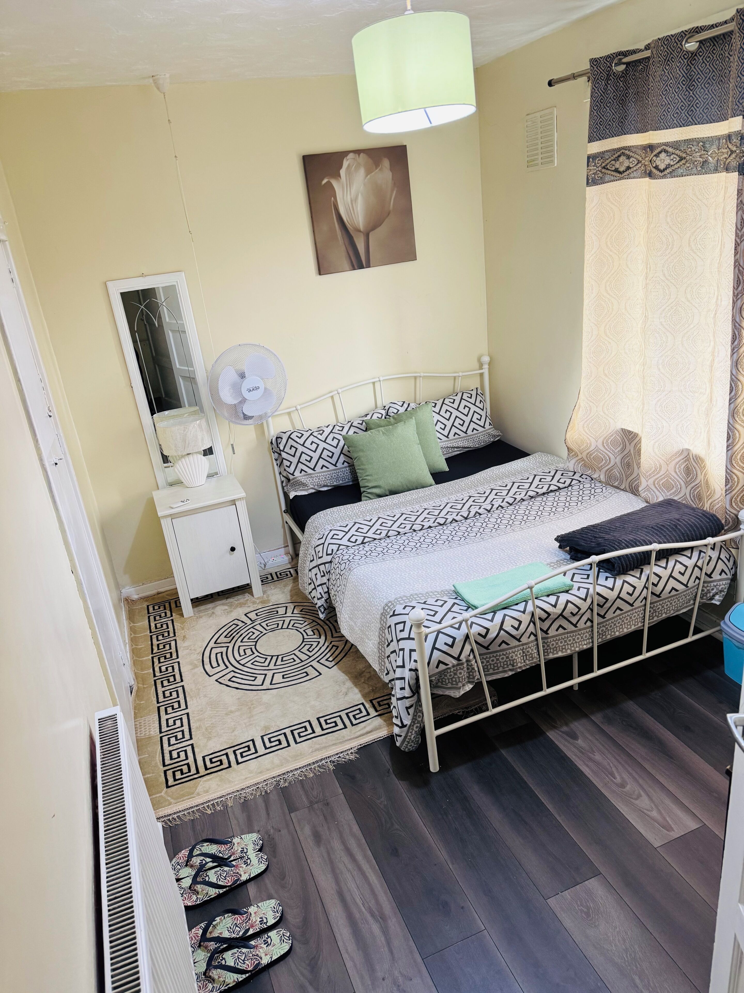 Een slaapkamer met een bed, nachtkastje, spiegel, ventilator en een kamerkleed met patroon.