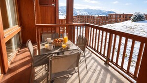 Un balcón de madera con una mesa preparada para el desayuno, con vistas a un paisaje montañoso nevado.