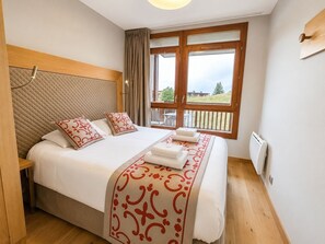 A hotel room with a bed, a window with a view, a wooden headboard, and a bedside table. - T4 au pied des pistes | Accès piscine | Balcon (Bourg-Saint-Maurice)